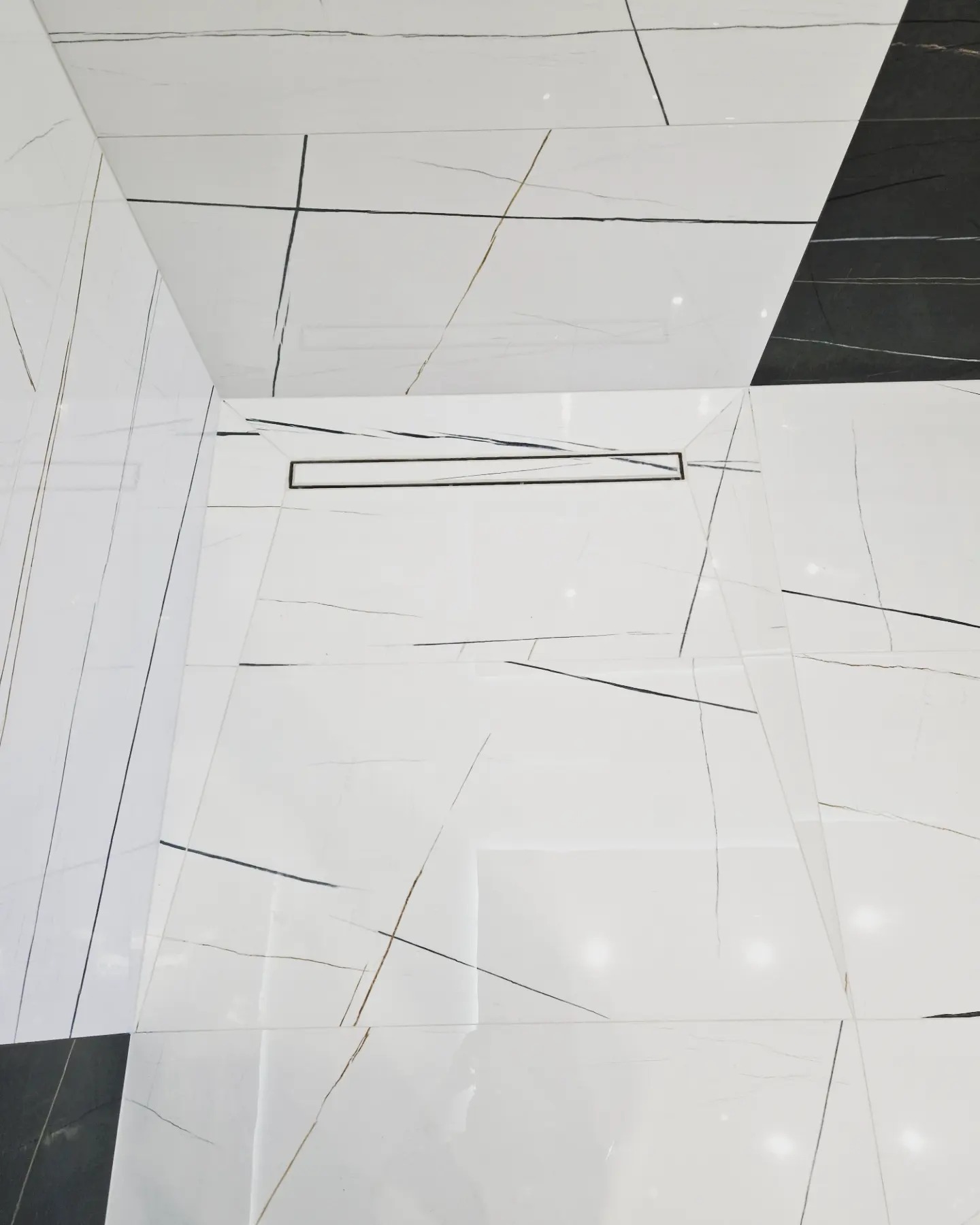 Białe i czarne płytki imitujące marmur z czarnymi żyłami, ułożone w nowoczesnej łazience z odpływem liniowym w podłodze.