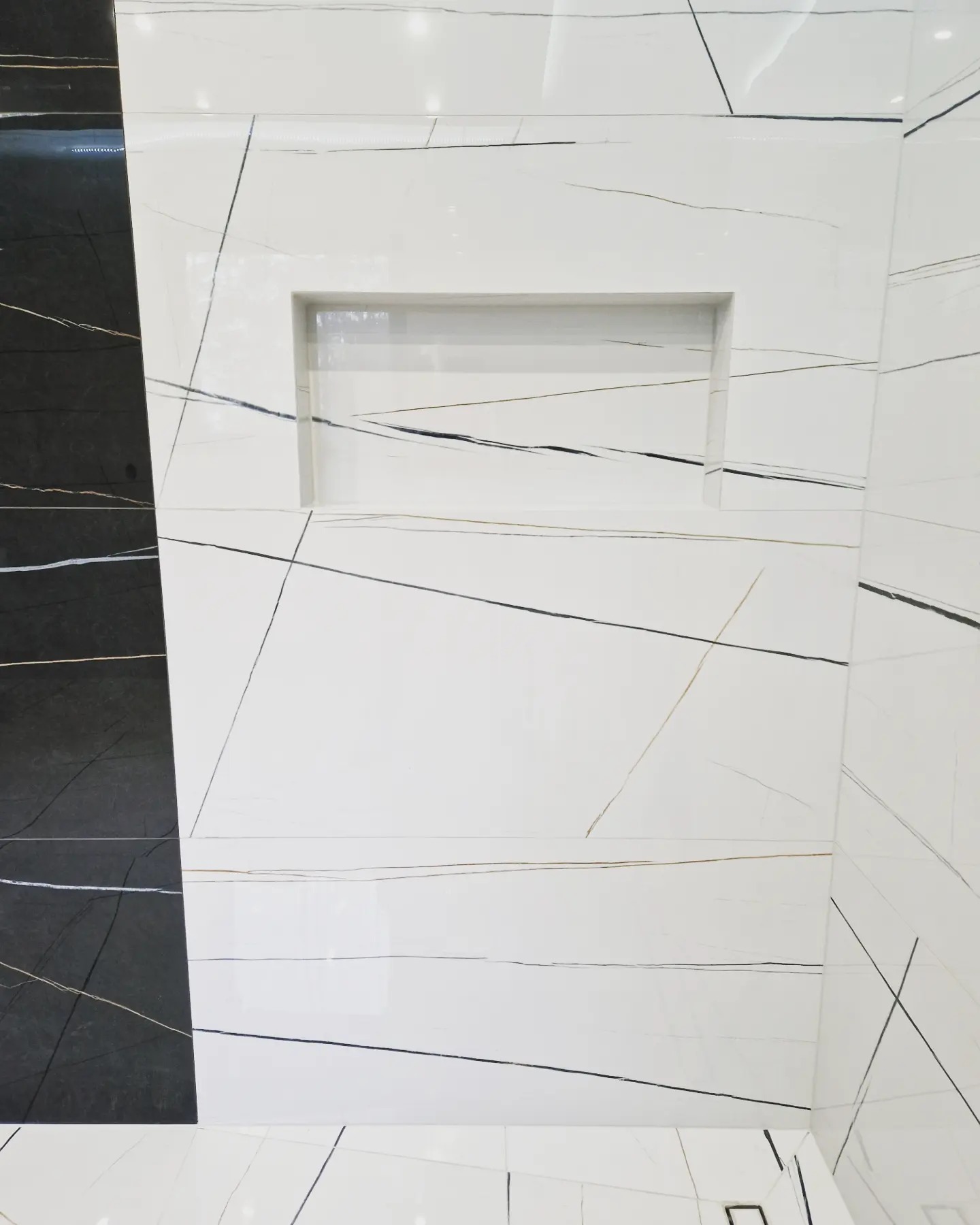 Ściana i podłoga w łazience wyłożone białymi płytkami imitującymi marmur z czarnymi i złotymi żyłami, z wnęką w ścianie, po lewej stronie fragment czarnej ściany z podobnym wzorem.