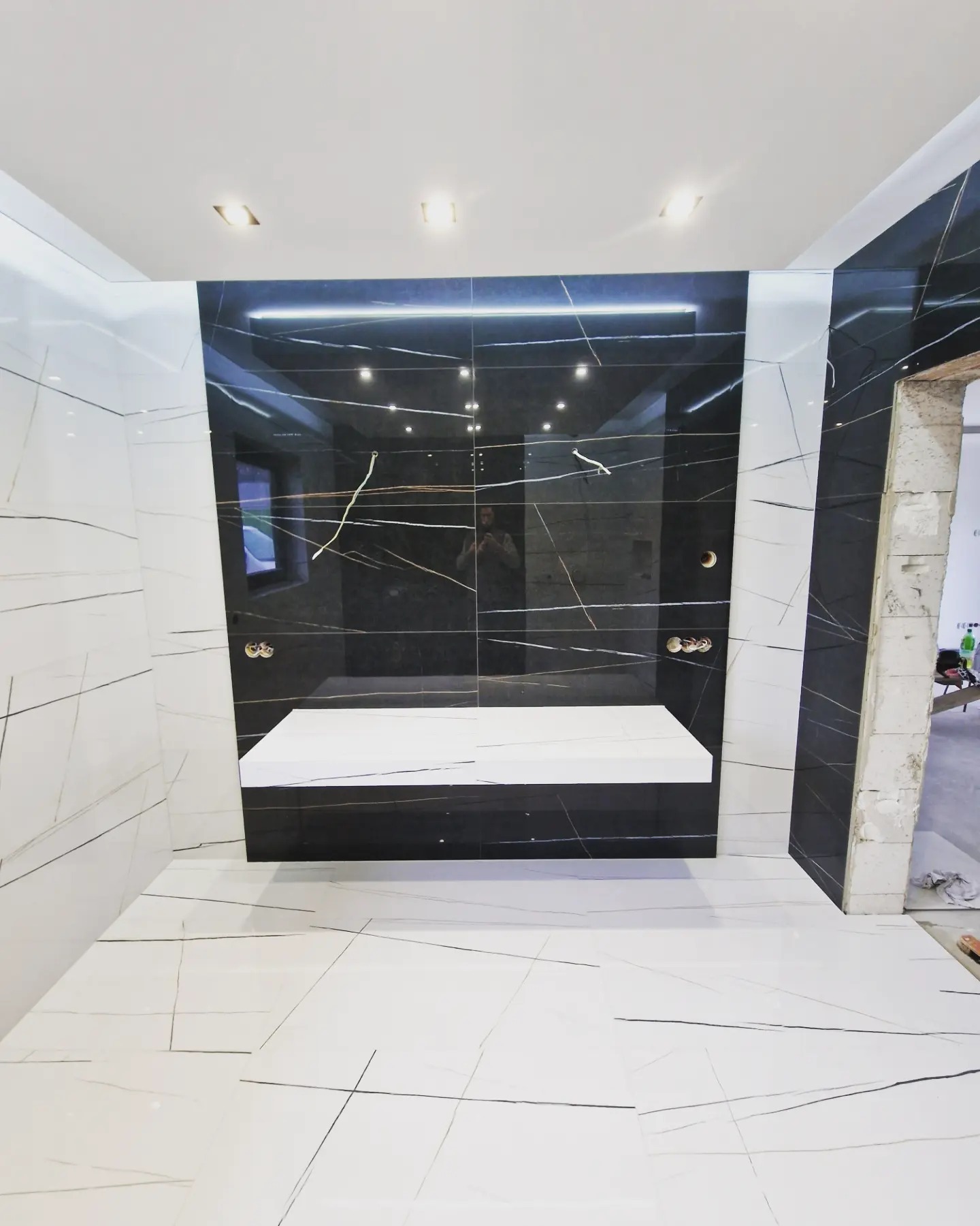 Nowoczesna łazienka z czarno-białymi płytkami imitującymi marmur, podświetlana półka pod umywalkę, widoczne instalacje wodne przed montażem armatury.