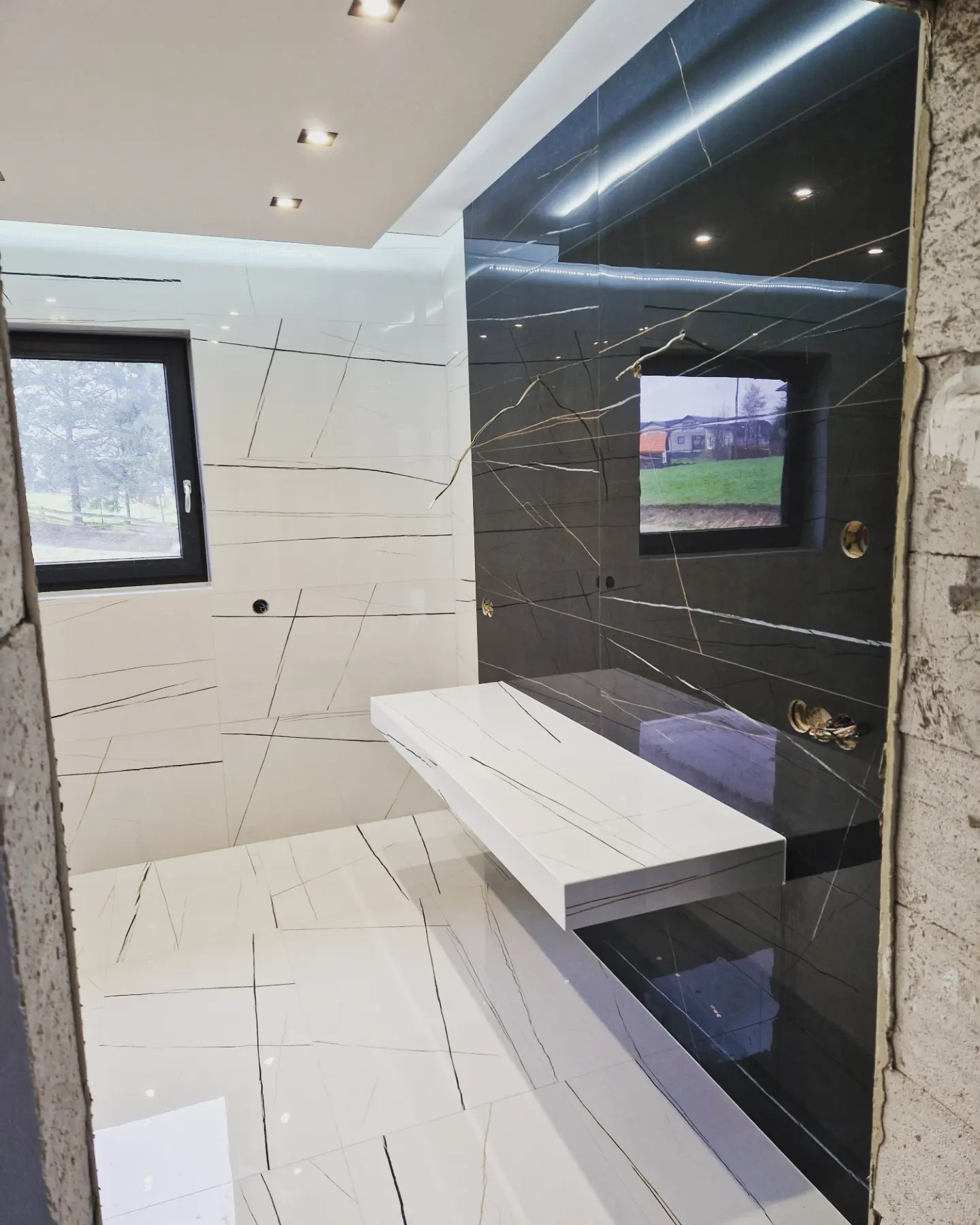 Nowoczesna łazienka w trakcie wykończenia z białymi i czarnymi płytkami imitującymi marmur, widoczne fragmenty niezabezpieczonych ścian, okno z widokiem na zewnątrz i minimalistyczna biała półka.
