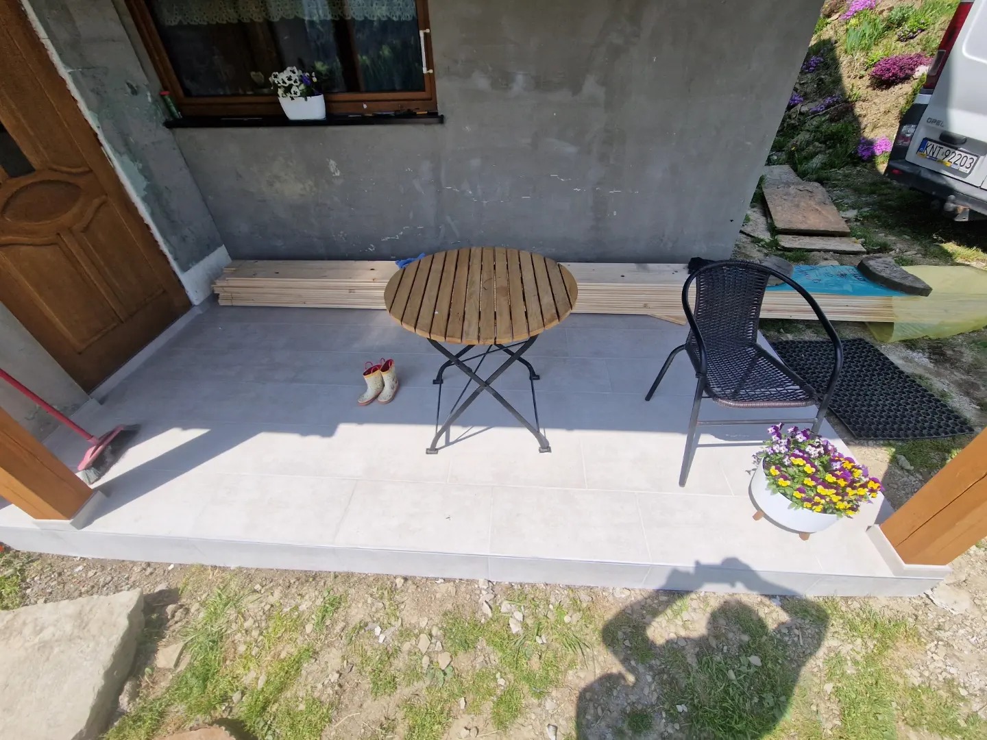 Wyłożony jasnymi płytkami taras przed domem z drewnianym stołem, krzesłem z wikliny i doniczką z bratkami; w tle ułożone deski i kalosze.