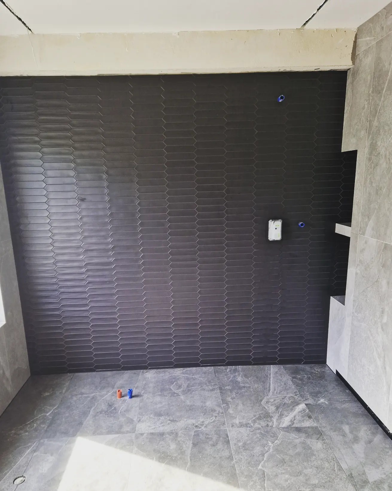 Ściana łazienki wyłożona ciemnoszarą, heksagonalną mozaiką ceramiczną, z widoczną instalacją elektryczną i fragmentem półki wnękowej, oraz podłoga z szarych płytek imitujących marmur, z niebieskim...