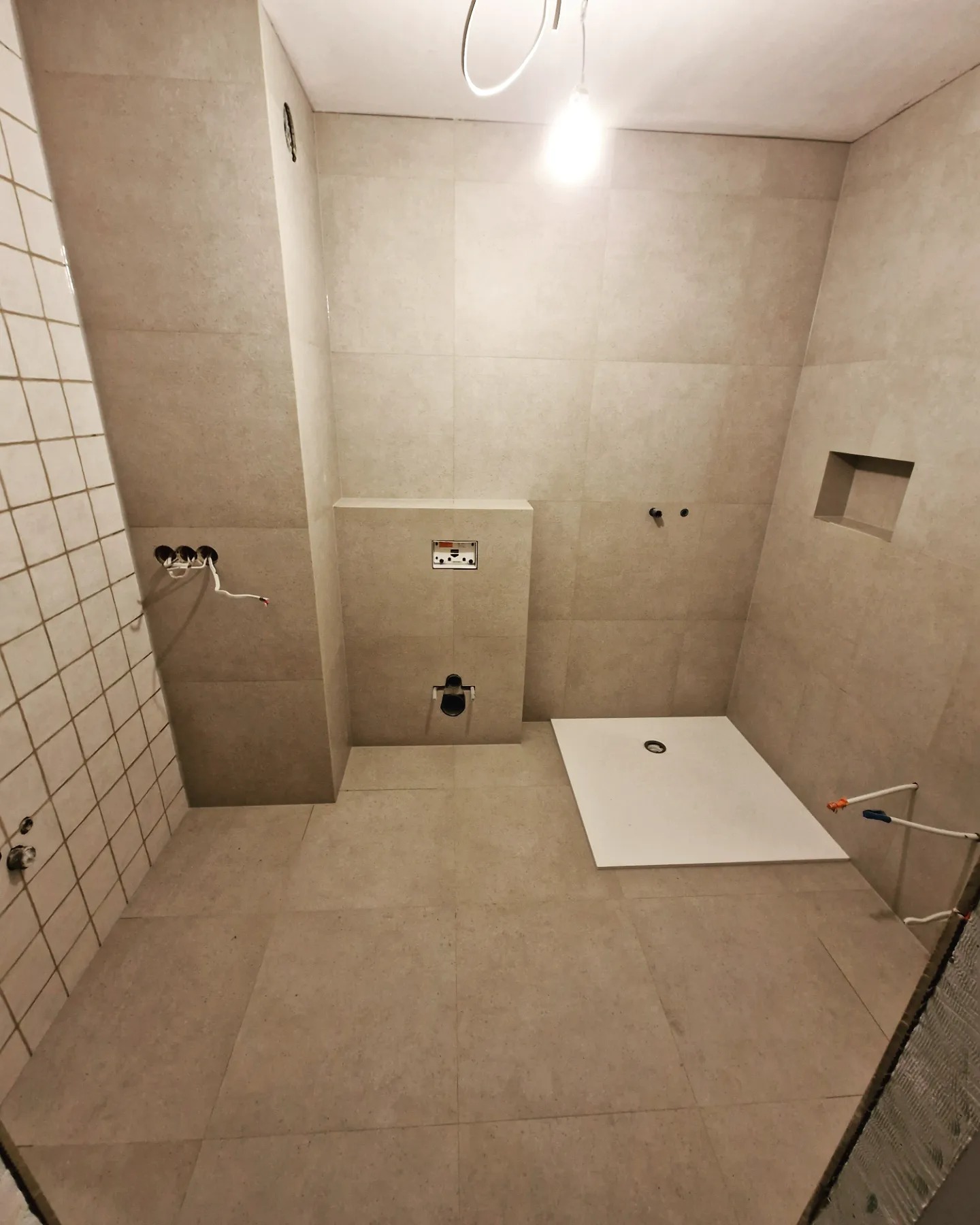 Wnętrze łazienki w trakcie wykańczania: częściowo ułożone beżowe płytki, biały brodzik, widoczne instalacje wodne i elektryczne, wnęka w ścianie.