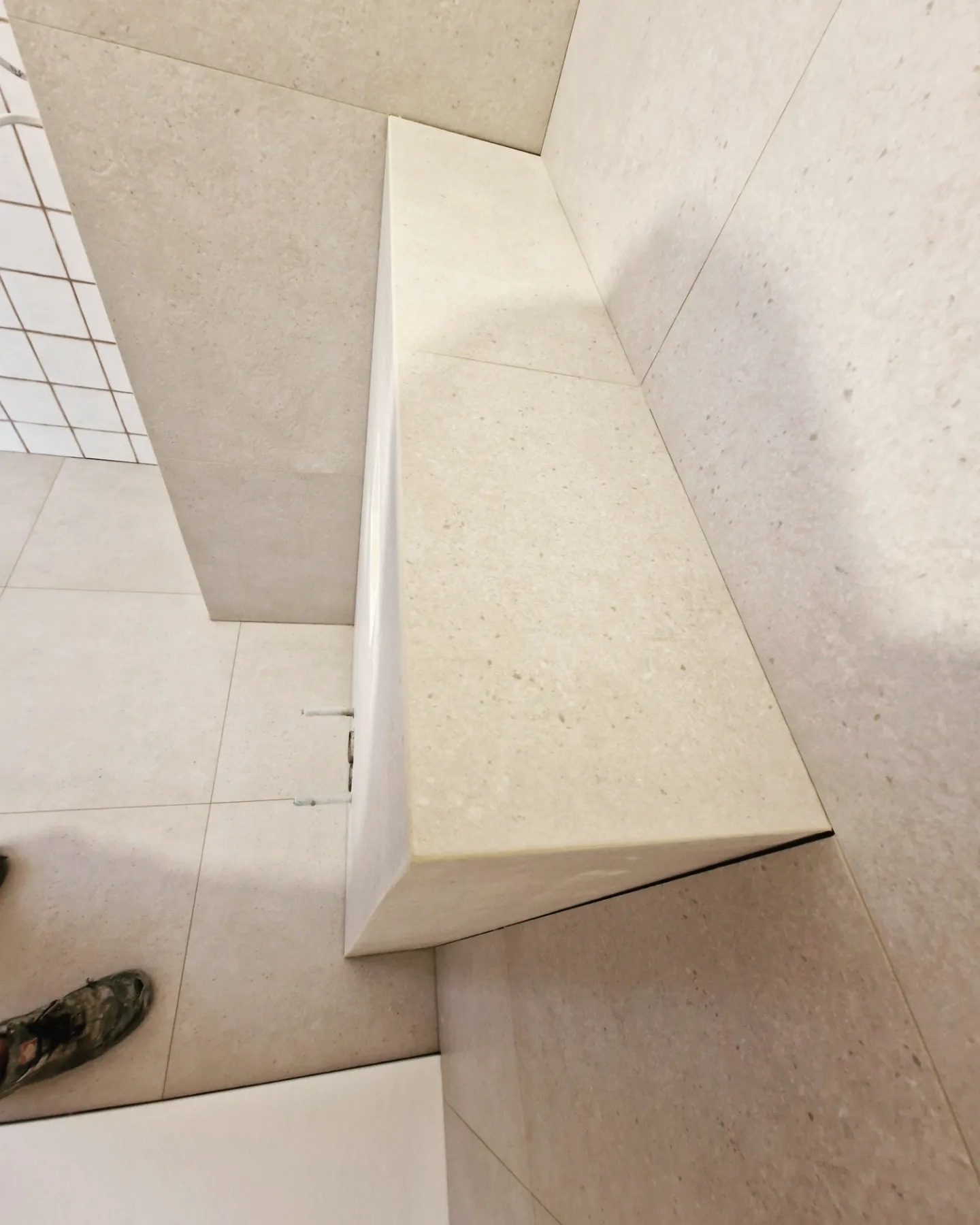 Fragment łazienki z wykończoną półką z płytek w kolorze beżowym, widoczne fugi i fragment podłogi z butem roboczym w lewym dolnym rogu.