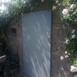 WALE Spółką z ograniczoną odpowiedzialnością - Szara, metalowa brama wejściowa do piwnicy lub pomieszczenia gospodarczego w murze z bloków, otoczona pnącymi różami.