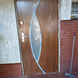 WALE Spółką z ograniczoną odpowiedzialnością - Brązowe drzwi wejściowe z owalnym przeszkleniem, zabezpieczone folią ochronną po montażu, widoczna pianka montażowa w ościeżnicy, obok ściana z cegły i miotła.