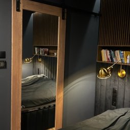 WALE Spółką z ograniczoną odpowiedzialnością - Drewniane drzwi przesuwne z czarnym okuciem w systemie szynowym, odbijające wnętrze sypialni z panelem ściennym, półką i lampką.