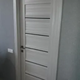 Zamknięte, białe drzwi wewnętrzne z poziomymi, czarnymi wstawkami, widoczne na tle szarej ściany z włącznikiem światła.