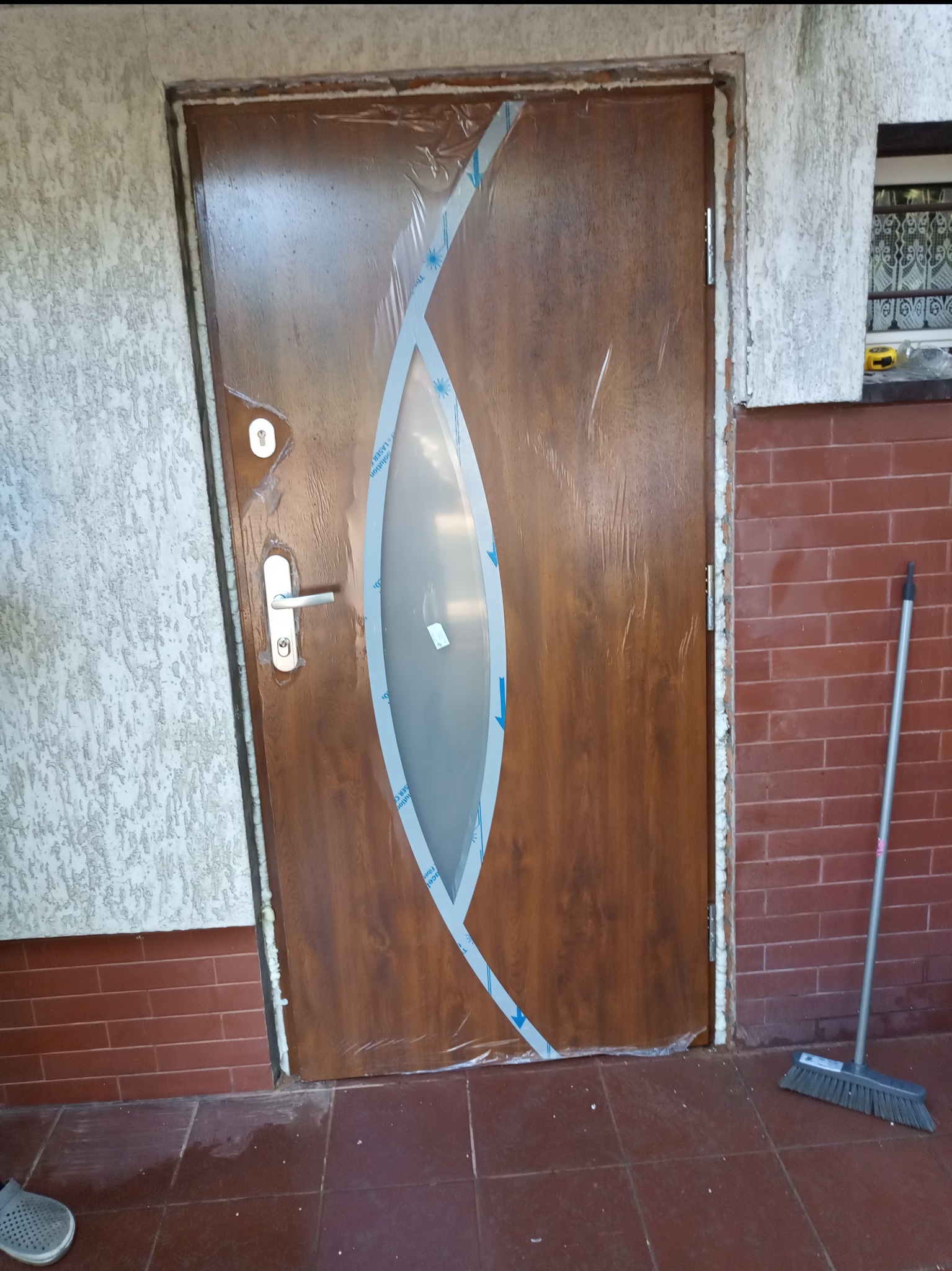 Brązowe drzwi wejściowe z owalnym przeszkleniem, zabezpieczone folią ochronną po montażu, widoczna pianka montażowa w ościeżnicy, obok ściana z cegły i miotła.