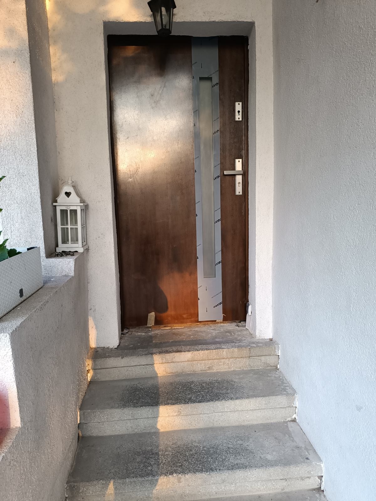 Brązowe drzwi wejściowe z częściowo usuniętą folią ochronną, zamontowane w białej elewacji z trzema betonowymi stopniami i ozdobnym lampionem obok.