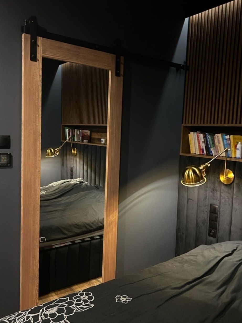 Drewniane drzwi przesuwne z czarnym okuciem w systemie szynowym, odbijające wnętrze sypialni z panelem ściennym, półką i lampką.