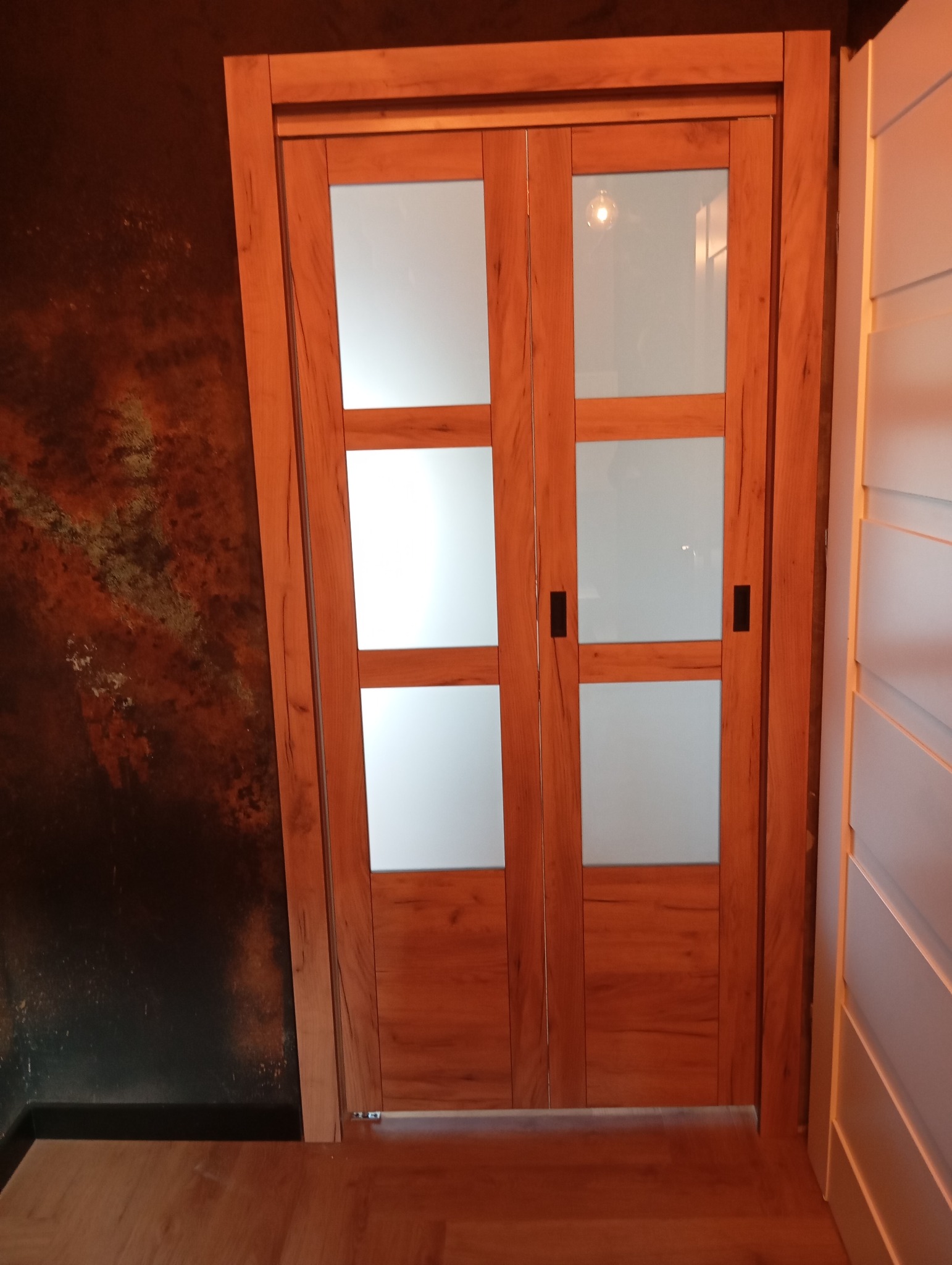Dwuskrzydłowe drzwi przesuwne z drewnianą ramą i matowym szkłem, zamontowane na tle ściany z efektem rdzy i białej szafy.