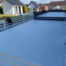 Biały taras z betonu z widocznymi stopniami, ogrodzeniem i zabudową w tle, wykonany w jasny, słoneczny dzień.