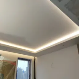 Nowoczesny, biały sufit z ukrytym oświetleniem LED, widoczny fragment okna i ściany w trakcie wykończenia.