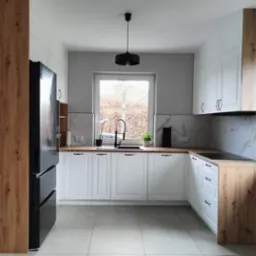 Nowoczesna kuchnia w bieli i drewnie: białe szafki dolne i górne kontrastują z drewnianymi blatami i wysoką zabudową, czarna lodówka i armatura, marmurowe panele nad blatem, minimalistyczna lampa...