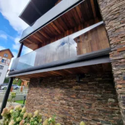 Trzy balkony z drewnianym wykończeniem i szklanymi balustradami, elewacja budynku wyłożona kamieniem, widok z dołu, w tle błękitne niebo z chmurami.