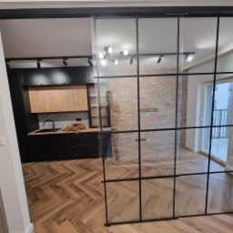 GlassView Ireneusz Turczyn - Szklane drzwi przesuwne z czarnymi szprosami oddzielają kuchnię od salonu. Parkiet w jodełkę, ceglana ściana w tle. Widoczny fragment zabudowy kuchennej.