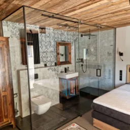 Nowoczesna łazienka z kabiną prysznicową oddzieloną szklaną ścianą, drewnianym sufitem i szafką pod umywalkę w stylu rustykalnym. Widoczny fragment sypialni.