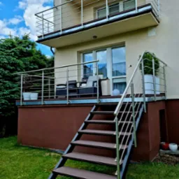 Dwupoziomowy taras z nierdzewną balustradą, drewnianymi schodami i zielonym trawnikiem. Na górnym balkonie antena. Nowoczesne wykończenie domu.