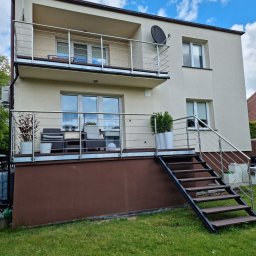 GlassView Ireneusz Turczyn - Dwukondygnacyjny dom z balkonami zabezpieczonymi nierdzewnymi balustradami. Schody z poręczą prowadzą na balkon. Na balkonach rośliny w donicach i meble.