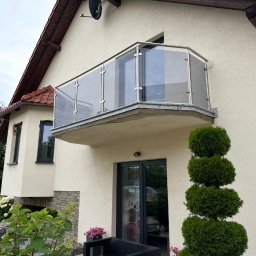 GlassView Ireneusz Turczyn - Szklana balustrada balkonowa z metalowymi słupkami, zamontowana na zewnątrz budynku z beżową elewacją. Widok z dołu, zieleń w tle.