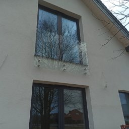 GlassView Ireneusz Turczyn - Elewacja domu z oknami aluminiowymi i szklaną balustradą balkonową mocowaną punktowo, w szybach odbija się drzewo i budynki.