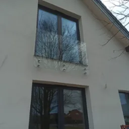 Elewacja domu z oknami aluminiowymi i szklaną balustradą balkonową mocowaną punktowo, w szybach odbija się drzewo i budynki.
