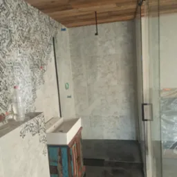 Nowoczesna łazienka w trakcie wykańczania: kabina prysznicowa ze szklanymi drzwiami, umywalka na szafce z postarzanego drewna, wzorzyste płytki na ścianie, drewniany sufit.