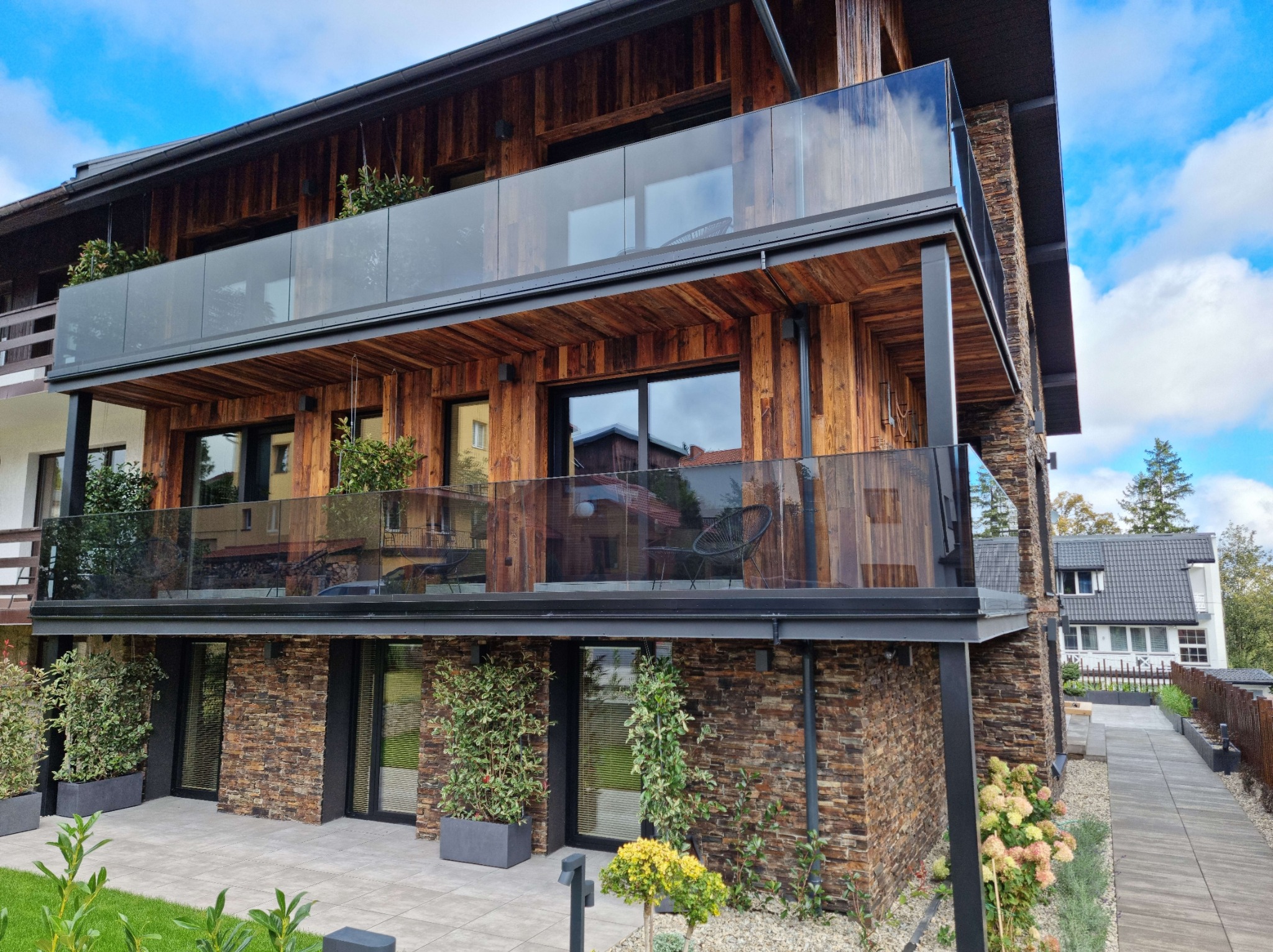 Nowoczesny budynek z drewnianą fasadą i szklanymi balustradami balkonowymi, widok z dołu, podkreślający architekturę i roślinność.