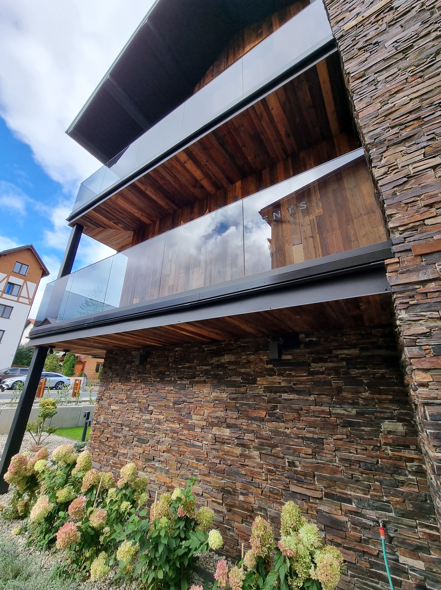 Trzy balkony z drewnianym wykończeniem i szklanymi balustradami, elewacja budynku wyłożona kamieniem, widok z dołu, w tle błękitne niebo z chmurami.