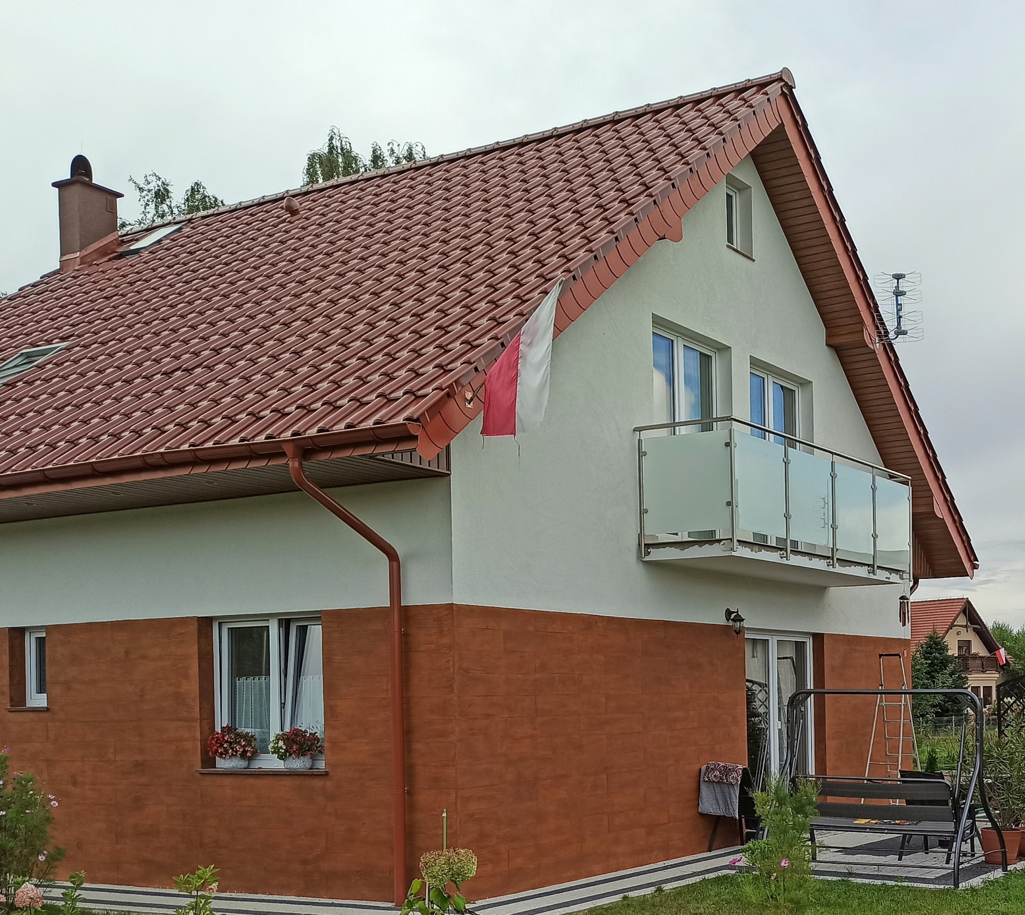Dom z balkonem zabezpieczonym szklaną balustradą, widok z zewnątrz, na dachu czerwona dachówka, w oknach kwiaty, powiewa flaga Polski.
