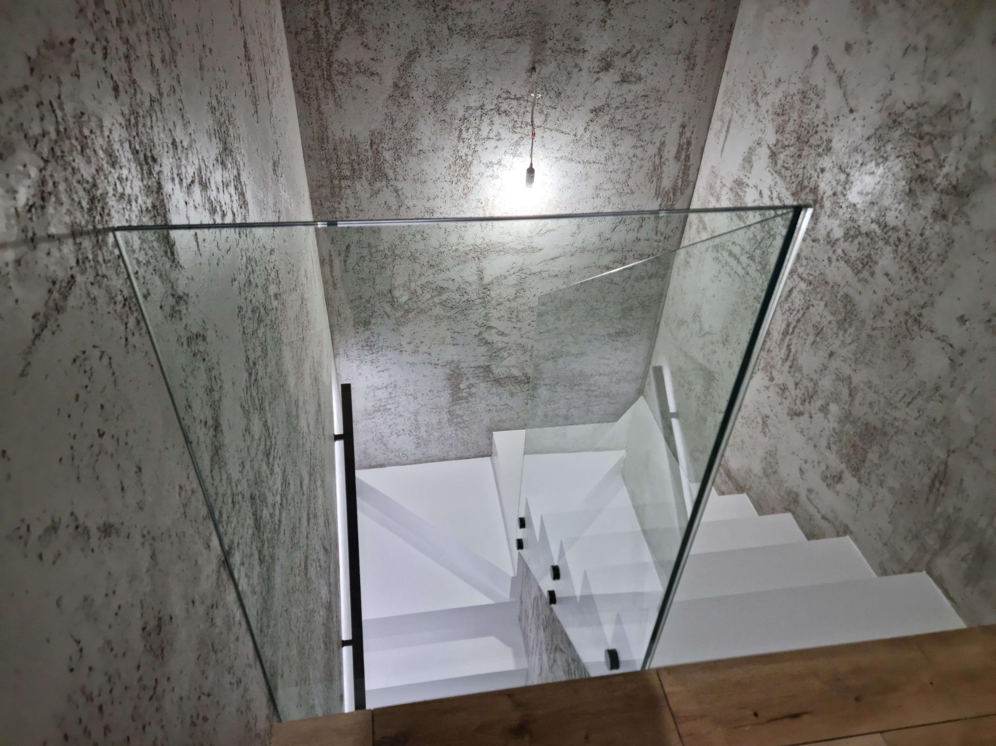 Szklana balustrada na białych schodach w surowym wnętrzu. Widok z góry na minimalistyczne rozwiązanie architektoniczne z elementami drewna w dolnej części kadru.