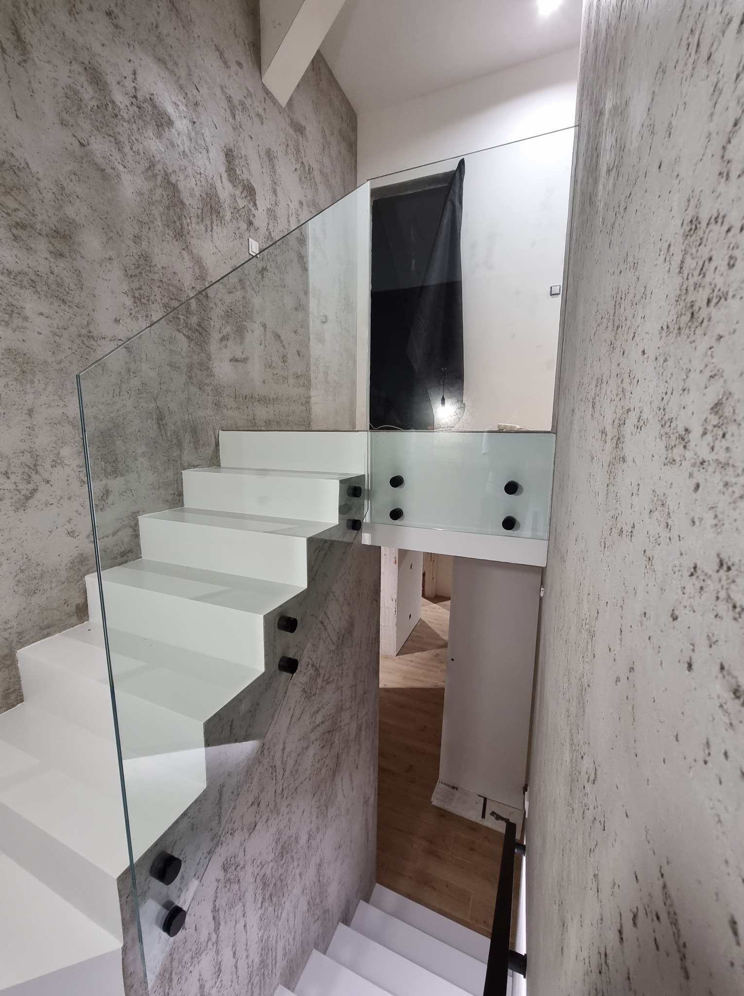 Nowoczesne, białe schody z balustradą ze szkła hartowanego, mocowaną na czarnych uchwytach. Ściana wykończona w stylu betonu architektonicznego w Dzbanowie.