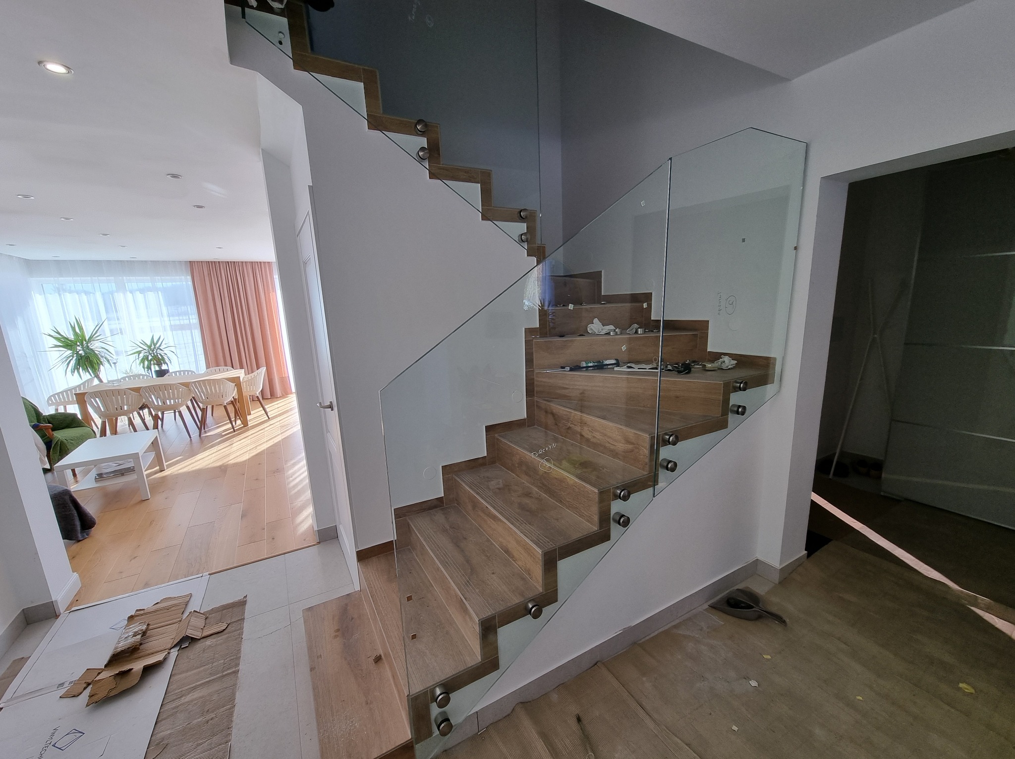 Nowoczesne schody drewniane z balustradą ze szkła bezramowego, zabezpieczającą przestrzeń. Jasne wnętrze z widoczną jadalnią w tle. Częściowo widoczne ślady montażu.