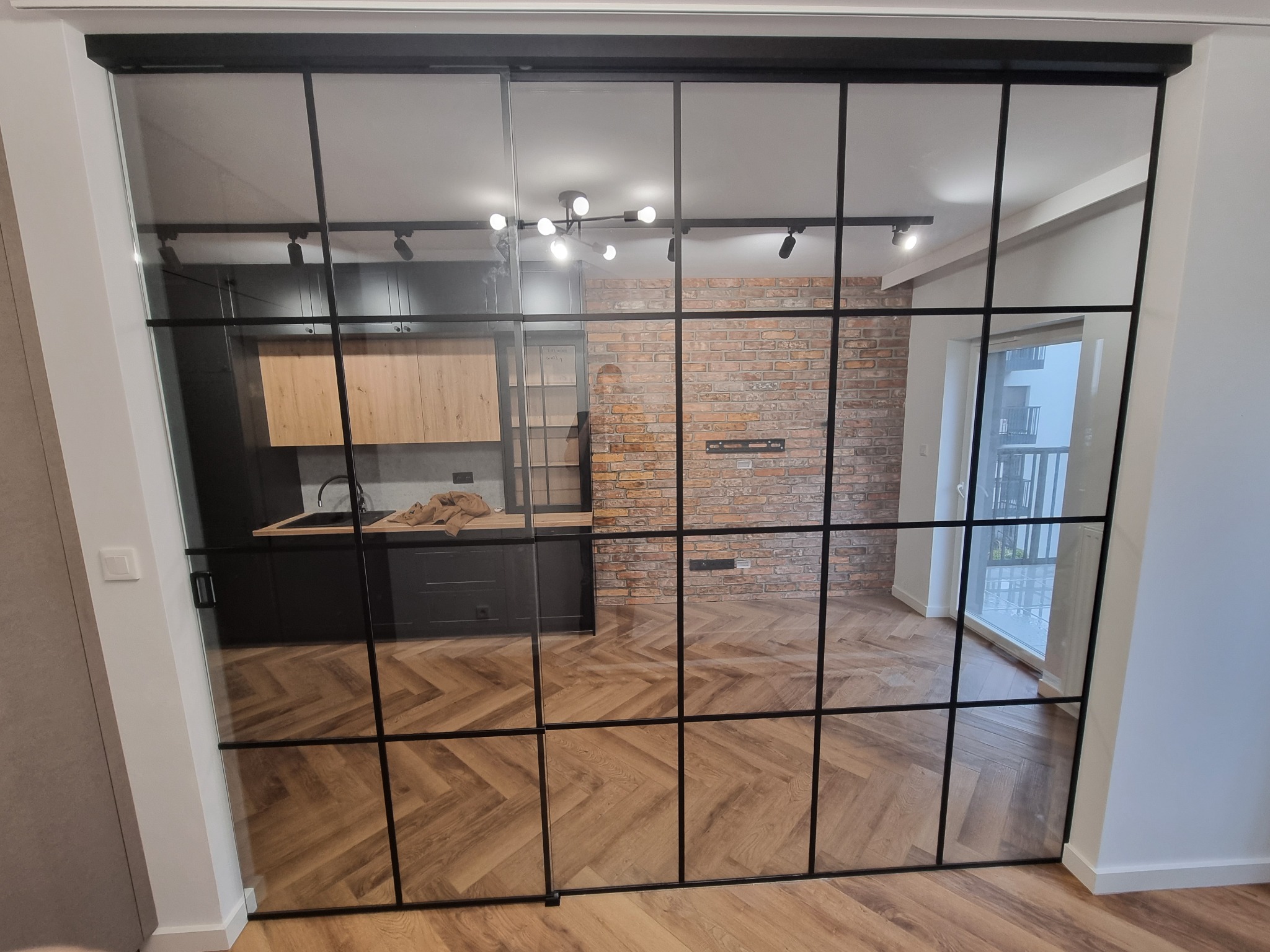 Szklane drzwi przesuwne z czarnymi szprosami oddzielają kuchnię od salonu. Widoczny parkiet w jodełkę i ceglana ściana w salonie. Nowoczesny design.