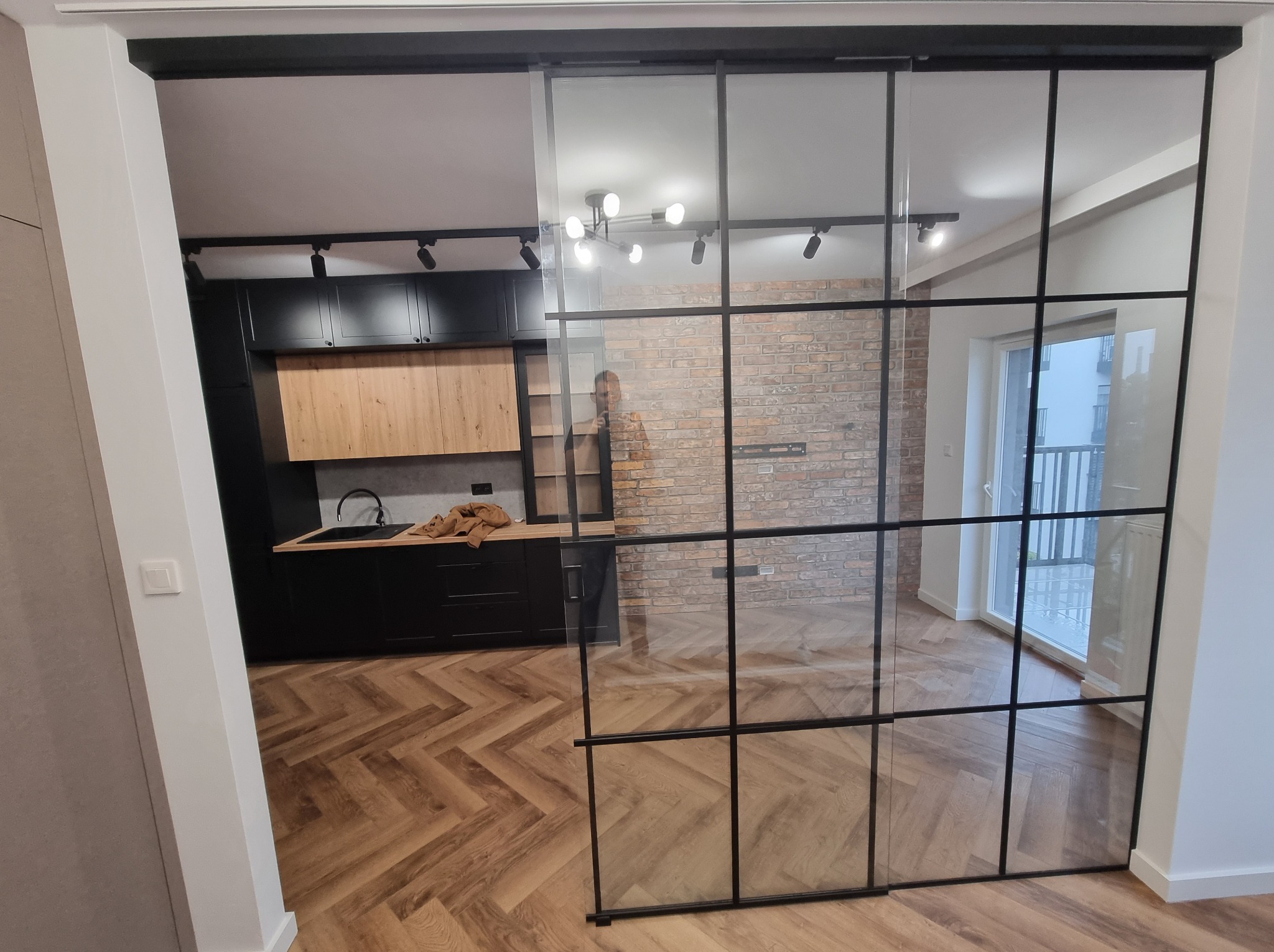 Szklane drzwi przesuwne z czarnymi szprosami oddzielają kuchnię od salonu. Parkiet w jodełkę, ceglana ściana w tle. Widoczny fragment zabudowy kuchennej.
