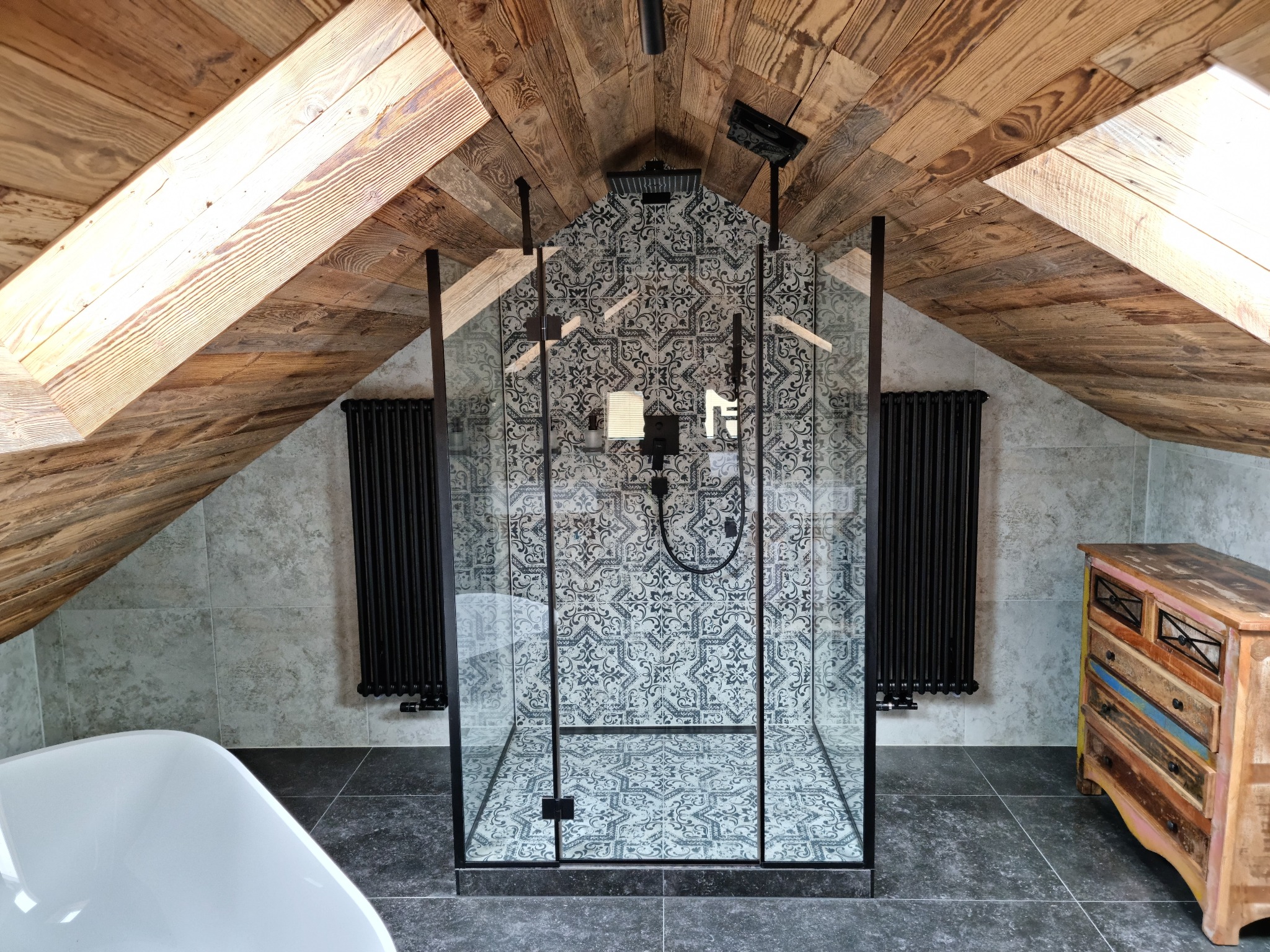 Szklana kabina prysznicowa z czarnymi ramami w łazience na poddaszu, drewniany sufit, wzorzyste płytki, grzejnik żeberkowy, komoda w stylu vintage.