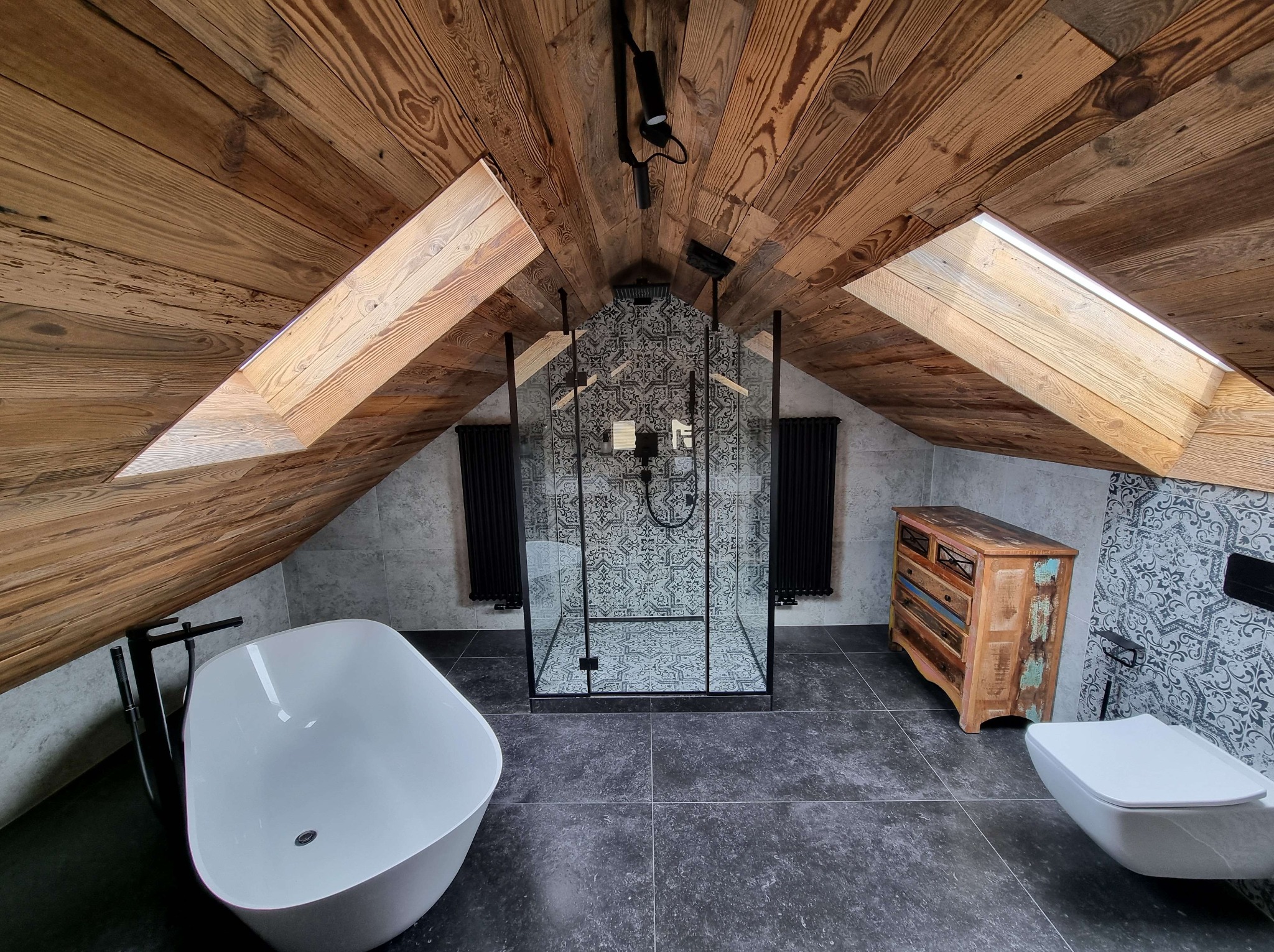 Aranżacja łazienki na poddaszu z drewnianym stropem, kabiną prysznicową ze wzorem, wolnostojącą wanną i komodą w stylu vintage. Widok z perspektywy żabiej.