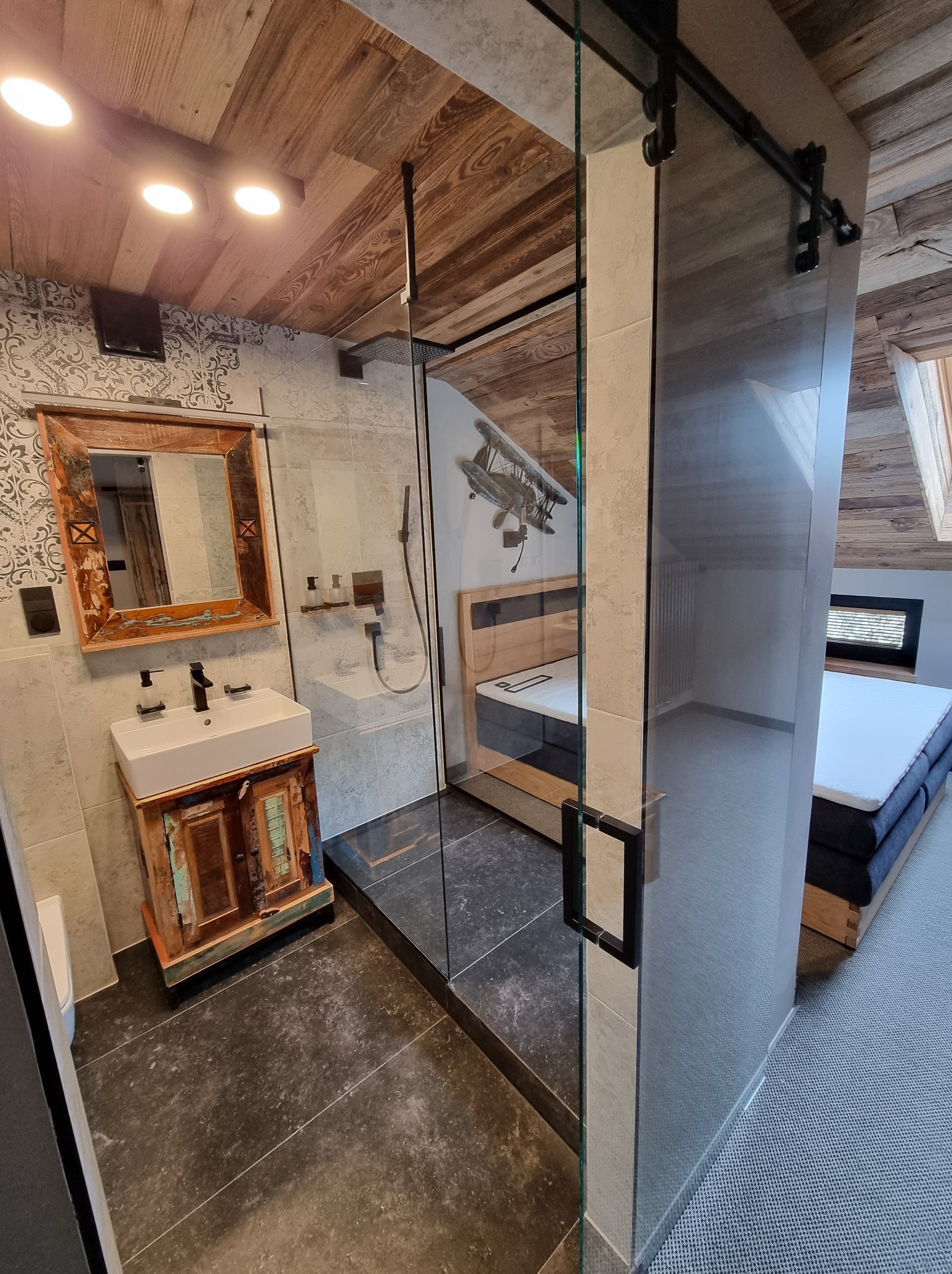 Nowoczesna łazienka z kabiną prysznicową ze szklanymi drzwiami przesuwnymi na szynie. Umywalka z szafką w stylu rustykalnym. Wykończenie w drewnie i szarościach.