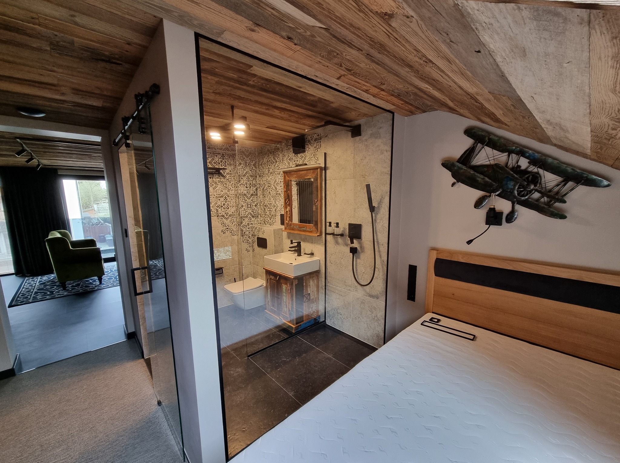 Nowoczesna łazienka za przesuwnymi drzwiami ze szkła, widok z sypialni. Ściany wykończone drewnem, dekoracja w postaci samolotu na ścianie. Widoczny fragment sofy.