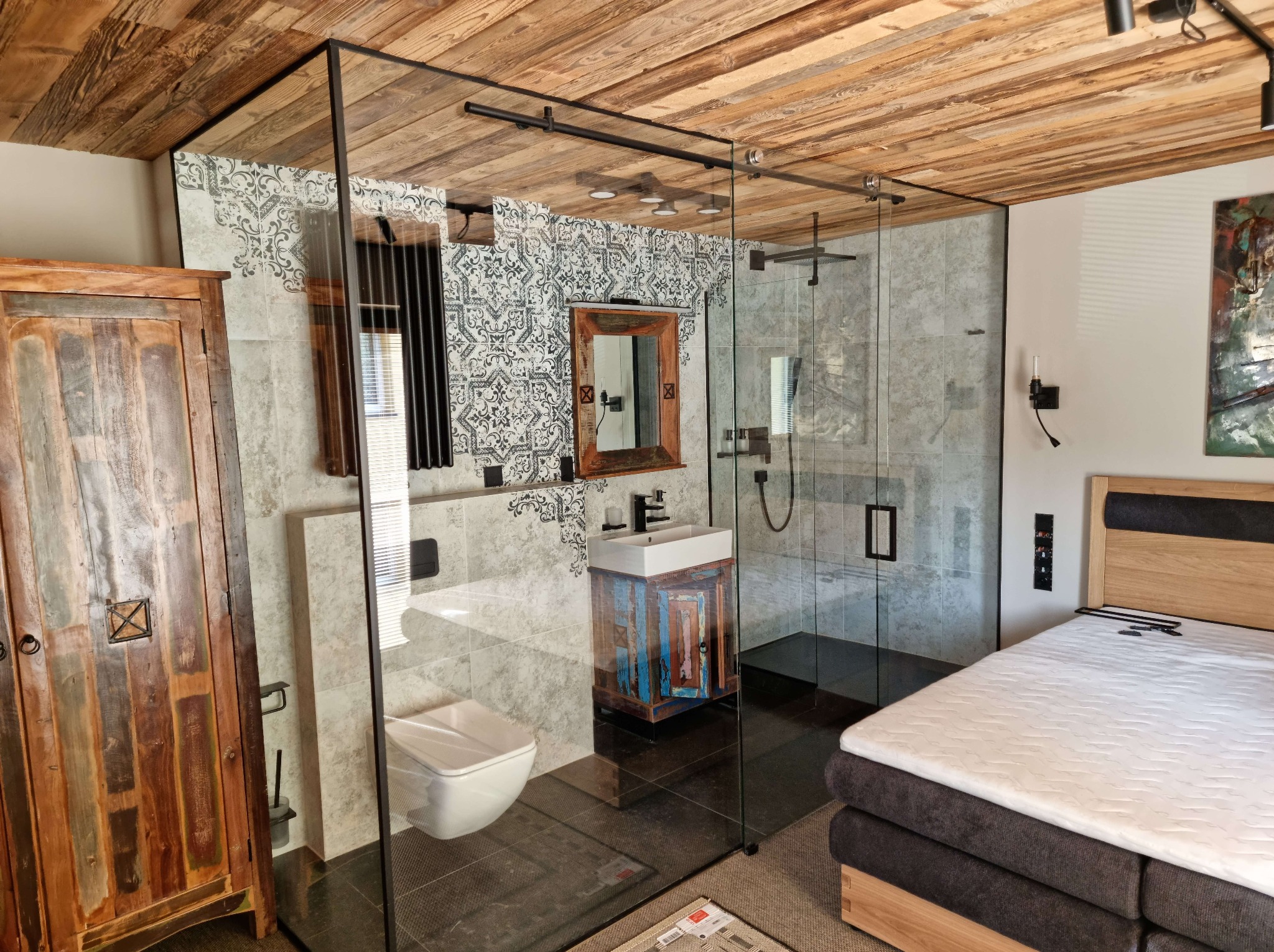 Nowoczesna łazienka z kabiną prysznicową oddzieloną szklaną ścianą, drewnianym sufitem i szafką pod umywalkę w stylu rustykalnym. Widoczny fragment sypialni.