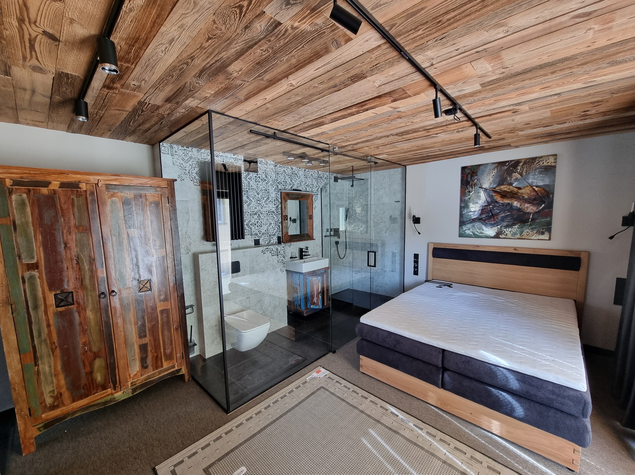 Nowoczesna łazienka z kabiną prysznicową ze szkła, umywalką i toaletą w pokoju z drewnianym sufitem. Widoczny fragment łóżka i szafy.