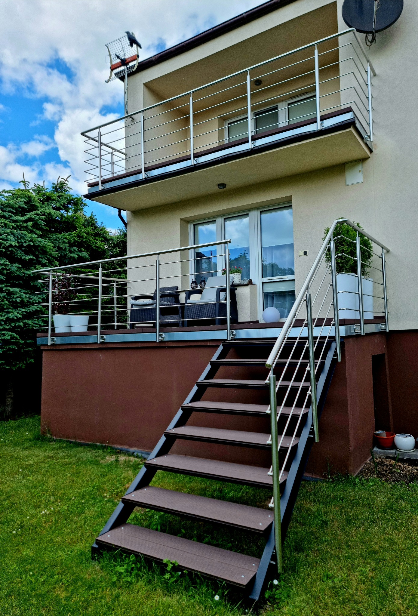 Dwupoziomowy taras z nierdzewną balustradą, drewnianymi schodami i zielonym trawnikiem. Na górnym balkonie antena. Nowoczesne wykończenie domu.