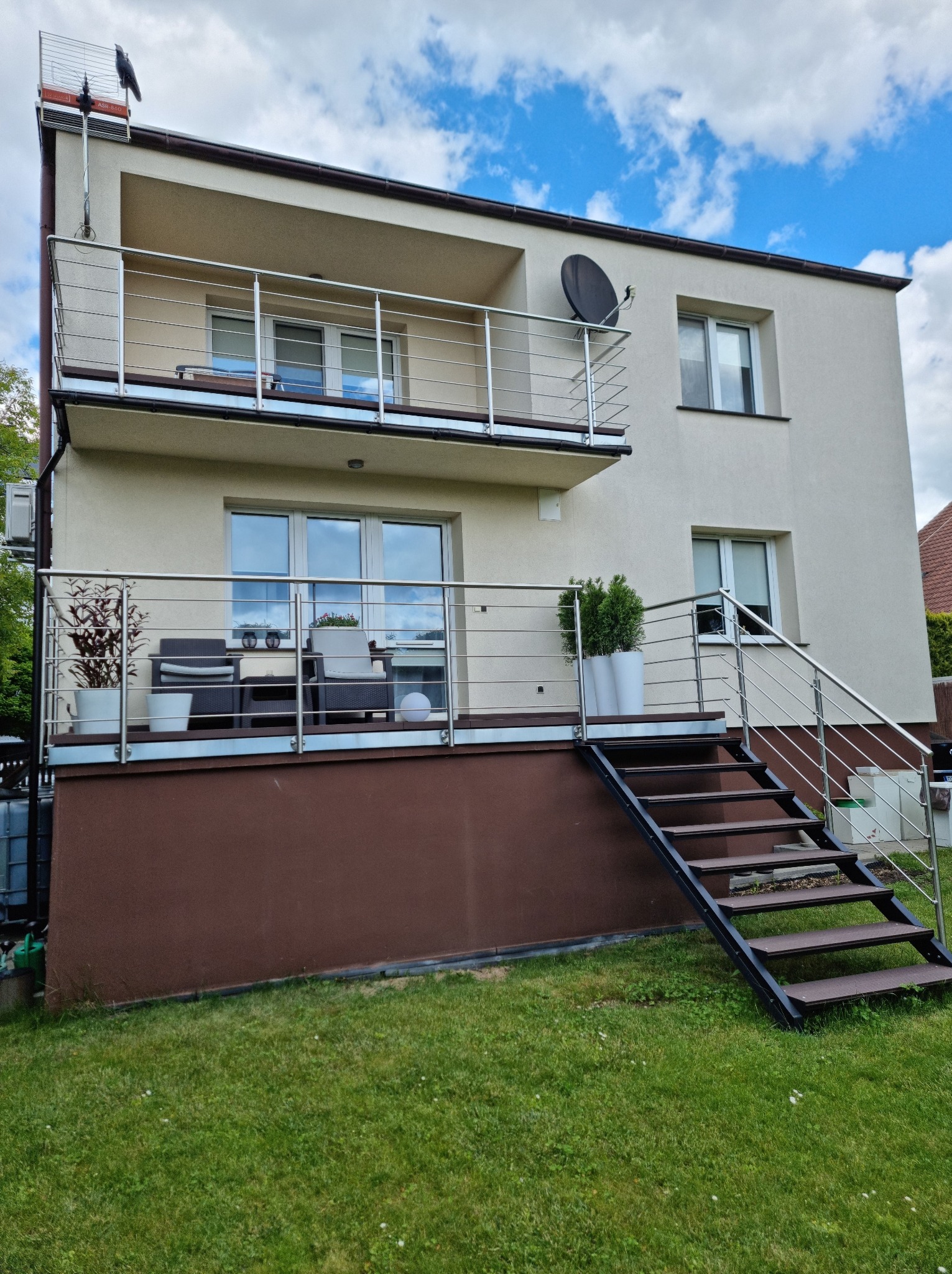 Dwukondygnacyjny dom z balkonami zabezpieczonymi nierdzewnymi balustradami. Schody z poręczą prowadzą na balkon. Na balkonach rośliny w donicach i meble.