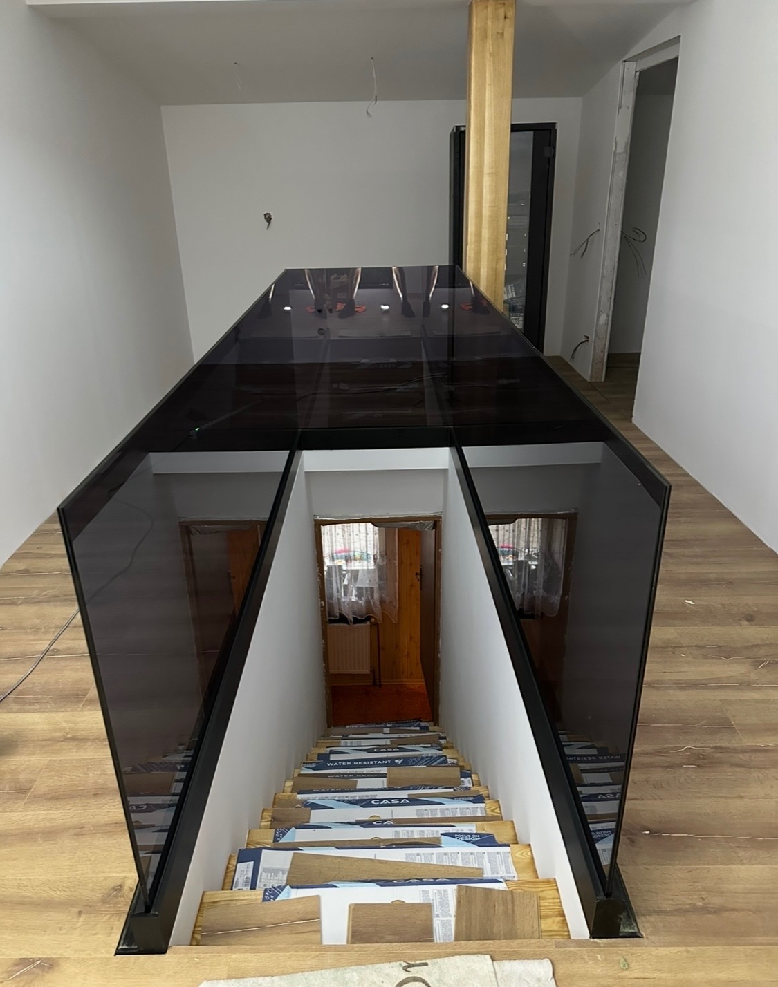 Szklana balustrada schodowa z czarnymi profilami w domu w Dzbanowie. Widok z góry na schody zabezpieczone kartonem podczas montażu.