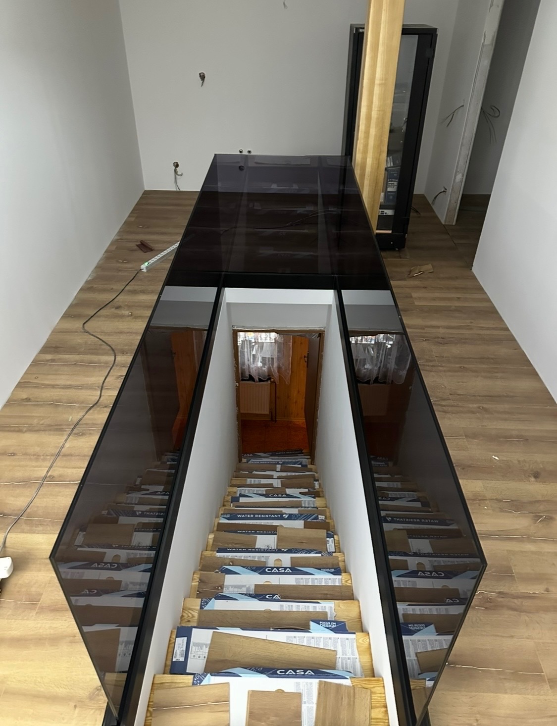 Nowoczesna szklana podłoga nad schodami, z widocznym odbiciem schodów w szkle. Schody zabezpieczone kartonami. Jasne ściany i podłoga z paneli.