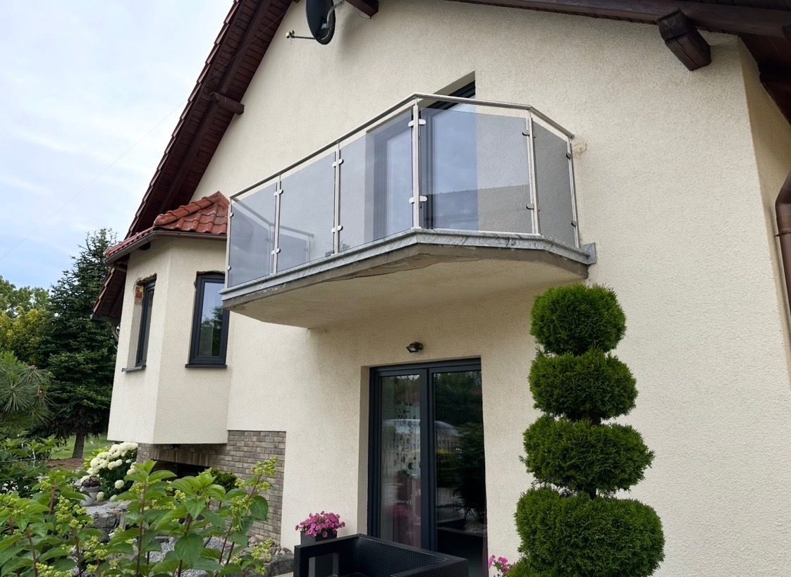 Szklana balustrada balkonowa z metalowymi słupkami, zamontowana na zewnątrz budynku z beżową elewacją. Widok z dołu, zieleń w tle.