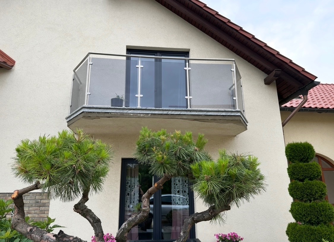 Balkon z przeszkloną balustradą i aluminiowymi oknami w nowoczesnym domu. Widok z dołu, z dekoracyjnymi drzewkami w pierwszym planie.