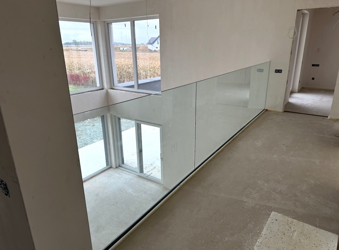Wnętrze domu w trakcie wykańczania z zamontowaną szklaną balustradą na antresoli. Widoczne duże okna z widokiem na zewnątrz i surowy beton na podłodze.