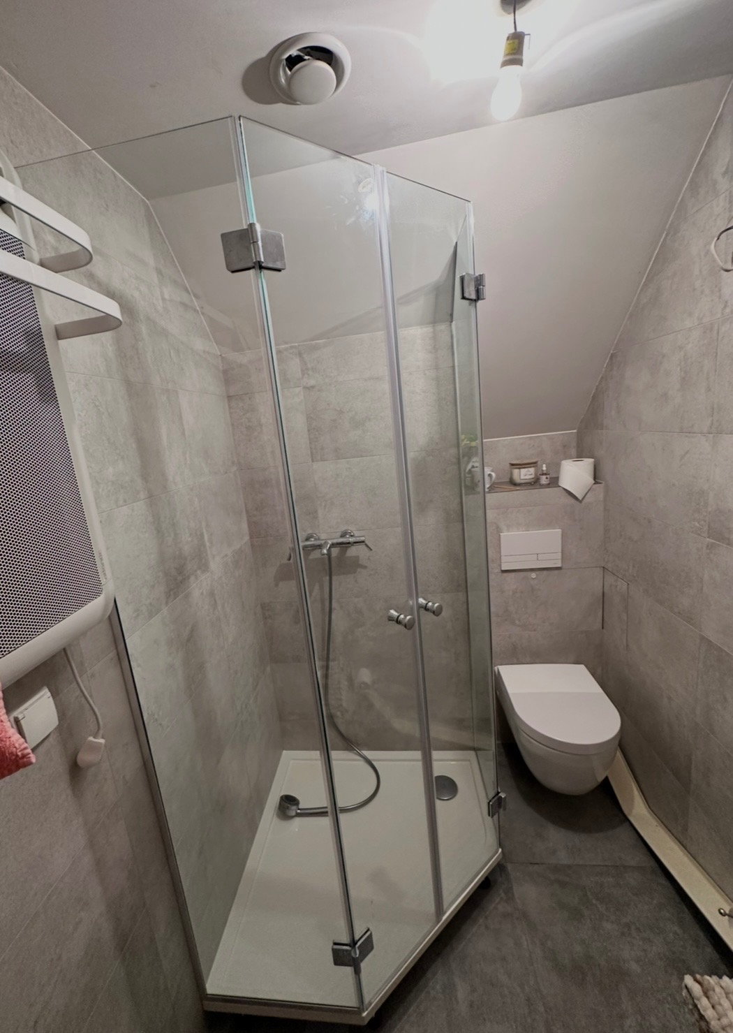 Szklana kabina prysznicowa w łazience z szarymi płytkami, widok na brodzik i baterię. Uwagę zwraca nietypowy kształt kabiny dostosowany do skosu dachu.